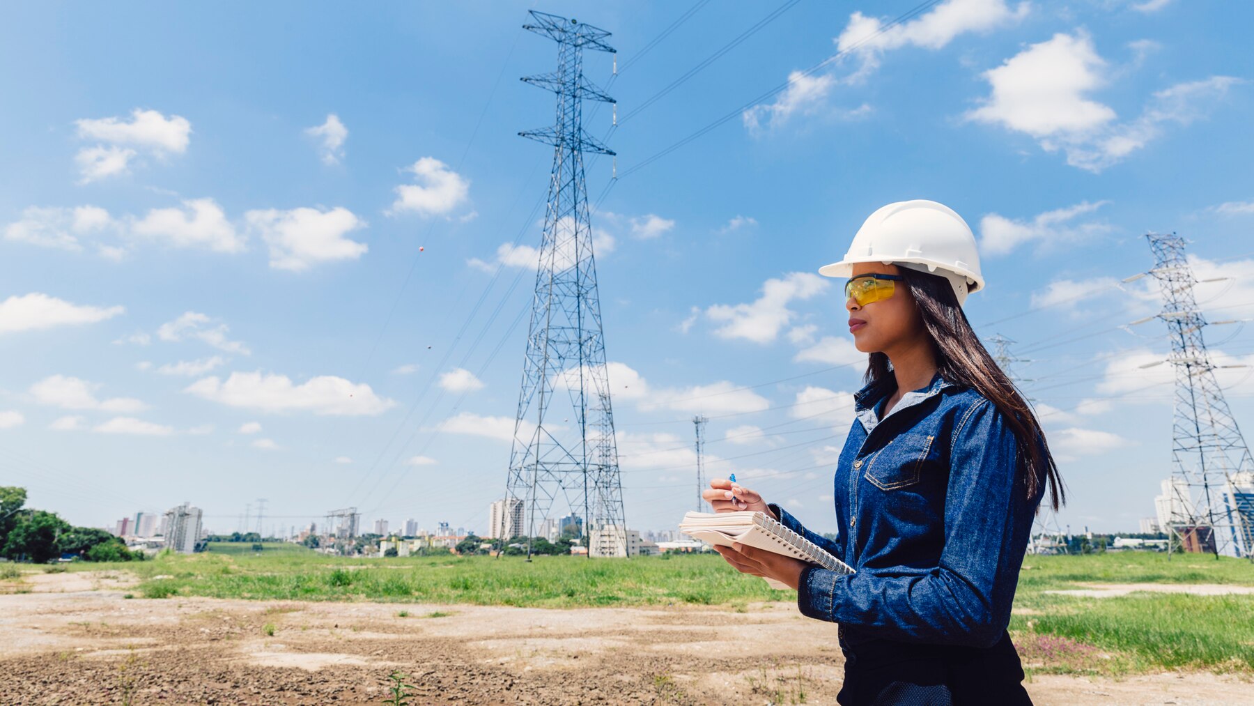 Female engineer wearing a safety helmet and glasses, taking notes near high-voltage power lines in an open field