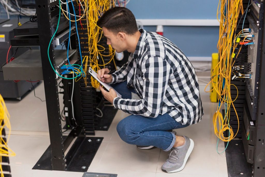 IT engineer inspecting server cables in a data center while taking notes on a clipboard.