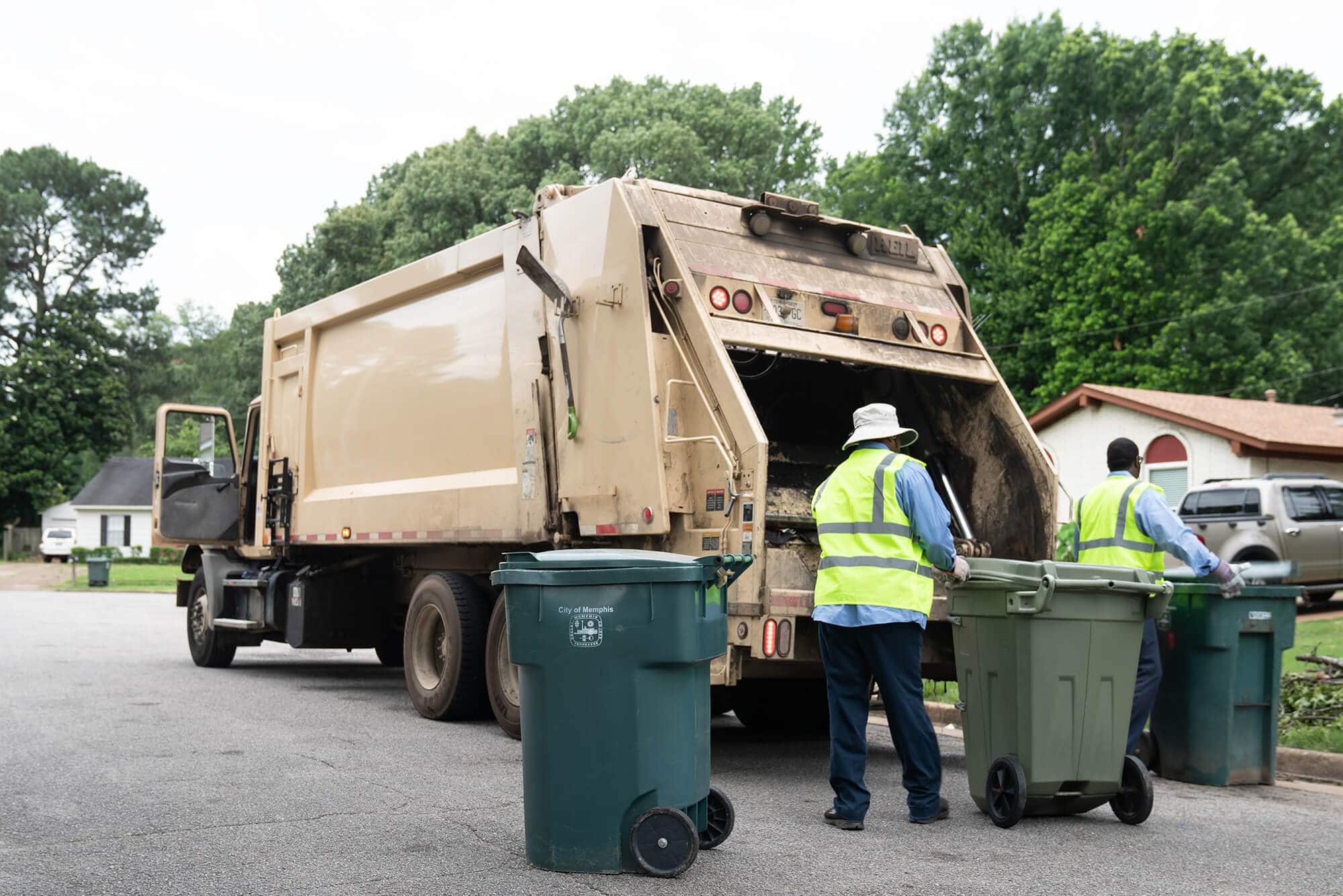 Two sanitation workers in reflective vests loading trash bins into a garbage truck on a residential street.